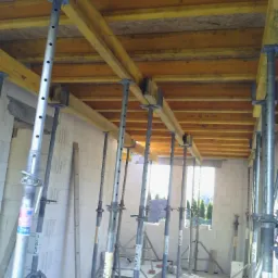 Wnętrze budynku w trakcie budowy z widocznym szalunkiem stropu podpartym metalowymi stemplami. Ściany z bloczków, okno z widokiem na zieleń.