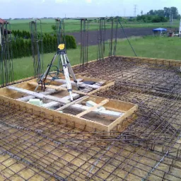 Zbrojenie fundamentu z użyciem metalowej siatki i drewnianych szalunków, poziomica na statywie, widok na zielone pola i zabudowę w tle, dzień.