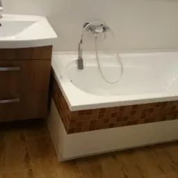 Nowoczesna łazienka: biała wanna z brązową mozaiką, umywalka z szafką w odcieniach drewna i armatura. Podłoga imitująca drewno.