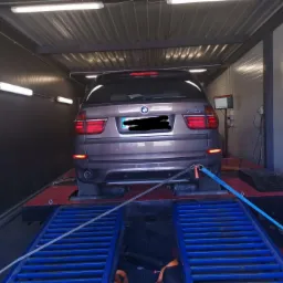 Brązowy samochód BMW X5 na hamowni podwoziowej, zabezpieczony pasami, w warsztacie z oświetleniem jarzeniowym.