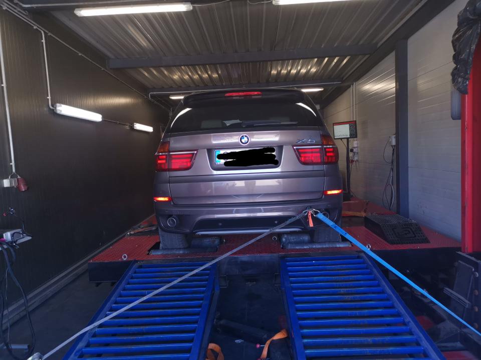 Brązowy samochód BMW X5 na hamowni podwoziowej, zabezpieczony pasami, w warsztacie z oświetleniem jarzeniowym.