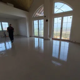 Świeżo wylana, gładka posadzka w jasnym pomieszczeniu z dużymi oknami, pracownik rozprowadza masę poziomującą, widoczne listwy dylatacyjne przy ścianach.