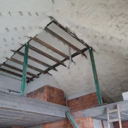 Poddasze z widoczną konstrukcją drewnianą i elementami mocującymi, pokryte białą pianą izolacyjną, widok z dołu w trakcie prac budowlanych.