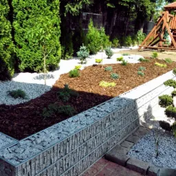 Nowoczesny ogród z gabionowym murkiem oporowym, korą, białym żwirem i różnorodną roślinnością. W tle drewniany plac zabaw.