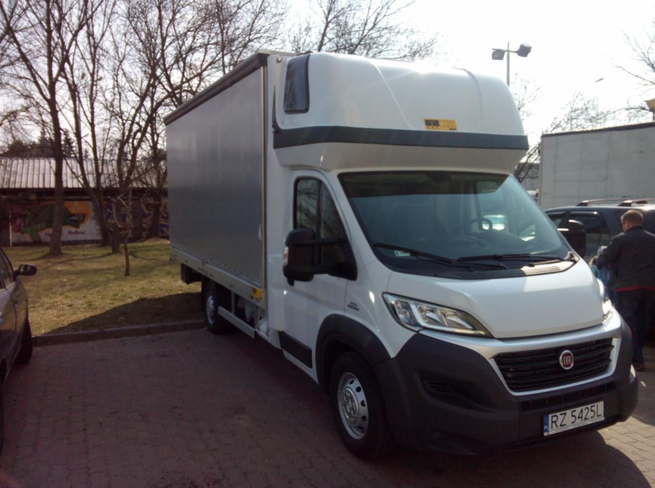 Biały bus dostawczy Fiat z szarą plandeką na parkingu, widok z boku na tle drzew i budynku z graffiti, tablica rejestracyjna RZ 5425L.
