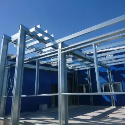 Montaż szkieletu hali stalowej na tle błękitnej ściany budynku, widoczne elementy mocujące i cień konstrukcji na posadzce.