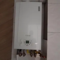 Biały, wiszący piec gazowy marki De Dietrich z podłączonymi rurami (miedzianymi, żółtymi i czarnymi) i zaworami, zamontowany na ścianie wyłożonej białymi płytkami.