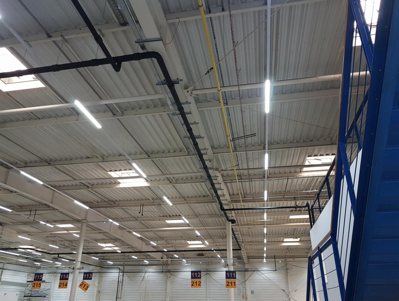 Rozległe wnętrze hali przemysłowej z widoczną instalacją rur pod sufitem, oświetleniem LED i fragmentem niebieskiej konstrukcji schodów.