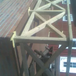 Drewniana konstrukcja balkonu z elementami w kształcie litery X, zabezpieczona taśmą malarską, przygotowana do wykończenia na tle ceglanego dachu.