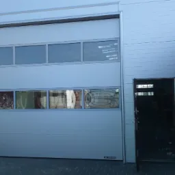 Zewnętrzna brama segmentowa w kolorze jasnoszarym z oknami, zamontowana w budynku o panelowej elewacji, obok otwarte drzwi wejściowe.