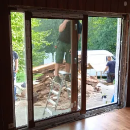 Demontaż starego okna: pracownicy usuwają ramę okienną, widoczny stos gruzu i elementów stolarki okiennej, drabina aluminiowa, przygotowanie do montażu nowej konstrukcji.