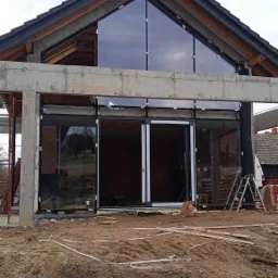 Surowy stan budynku z zamontowanymi dużymi, przesuwnymi oknami aluminiowymi i trójkątnym oknem szczytowym; widoczne betonowe elementy konstrukcyjne i ziemia na pierwszym planie.