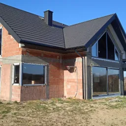 Surowy stan domu jednorodzinnego w trakcie budowy, widoczne okna z białymi ramami, ściany z czerwonej cegły, dach z czarnej dachówki, teren wokół domu jeszcze niezagospodarowany.