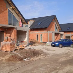 Wizualizacja budowy szeregu domów jednorodzinnych z czerwonej cegły, z czarnymi dachami, widoczne okna w szczytach, surowy stan, gruz, samochody na placu budowy.