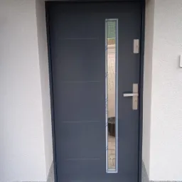 Grafitowe drzwi wejściowe z wąskim przeszkleniem, osadzone w ościeżnicy, widoczne ślady montażu na posadzce.