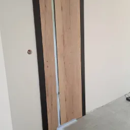 Zamknięte, przesuwne drzwi wewnętrzne z okleiną imitującą jasne drewno, obramowane czarną ramą, widoczne na tle białej ściany i szarej podłogi z płytek.