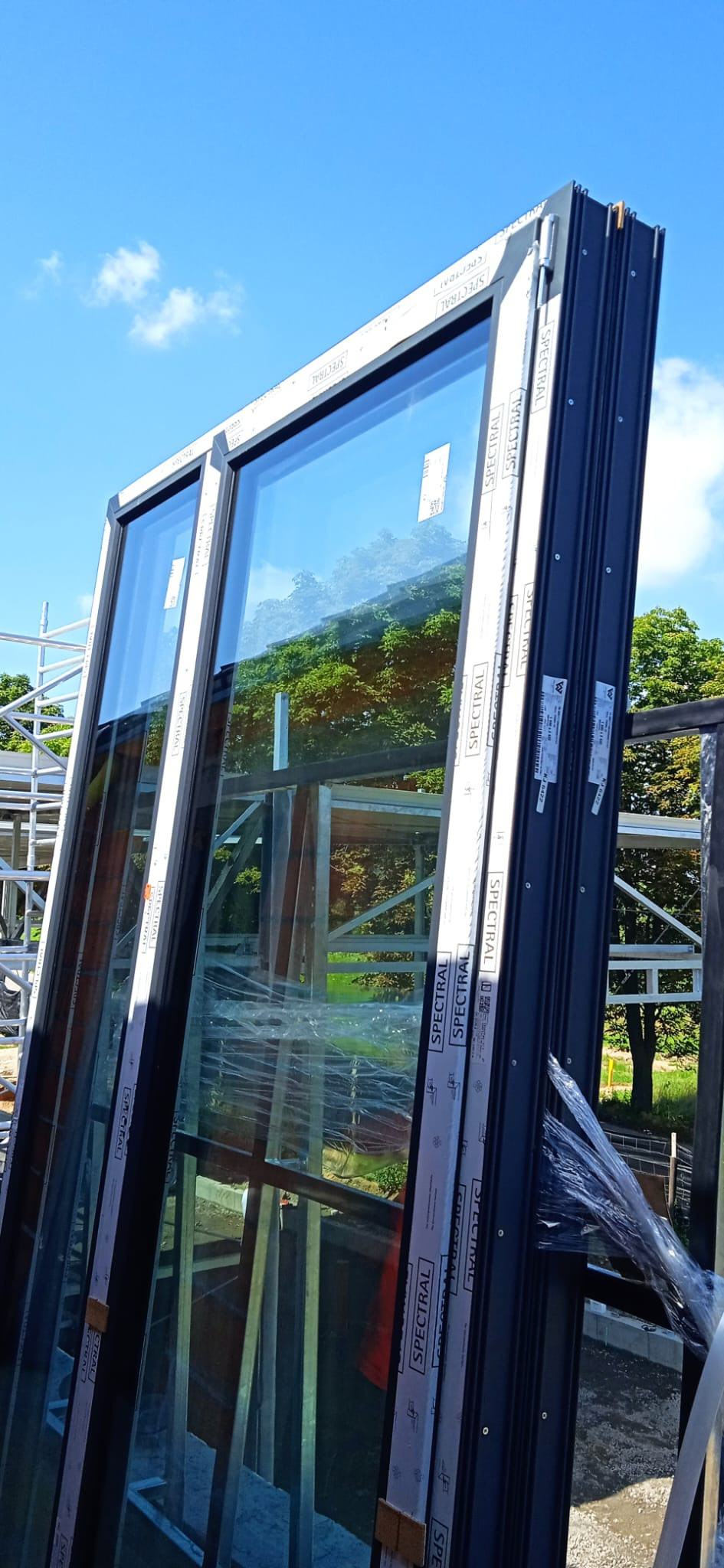 Nowe, duże okna z czarnymi ramami, zabezpieczone folią i taśmami ochronnymi z nadrukiem 'SPECTRAL', oparte na metalowej konstrukcji na zewnątrz budynku, z widocznym niebem i zielenią w tle.