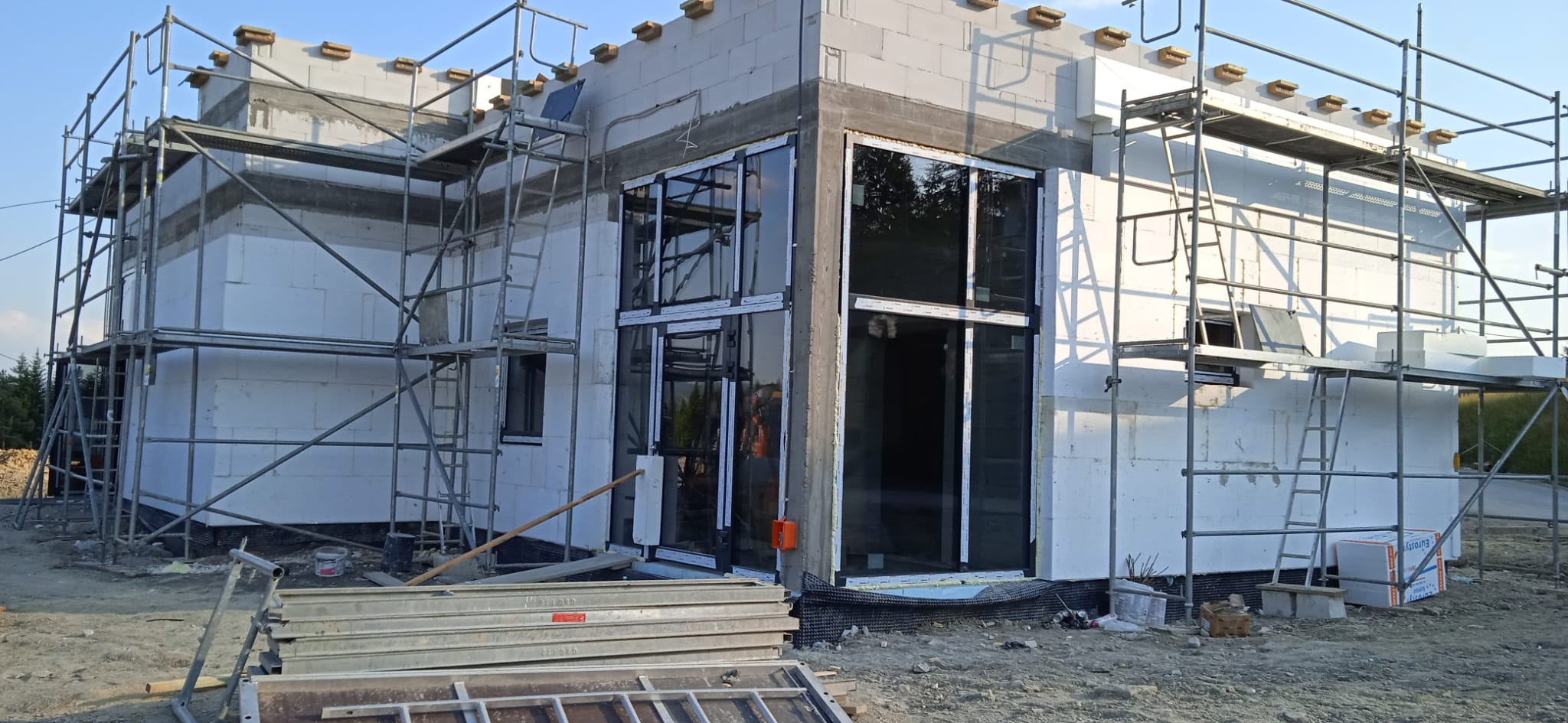 Budynek w trakcie budowy z zamontowanymi oknami i drzwiami, widoczne rusztowania i materiały budowlane.