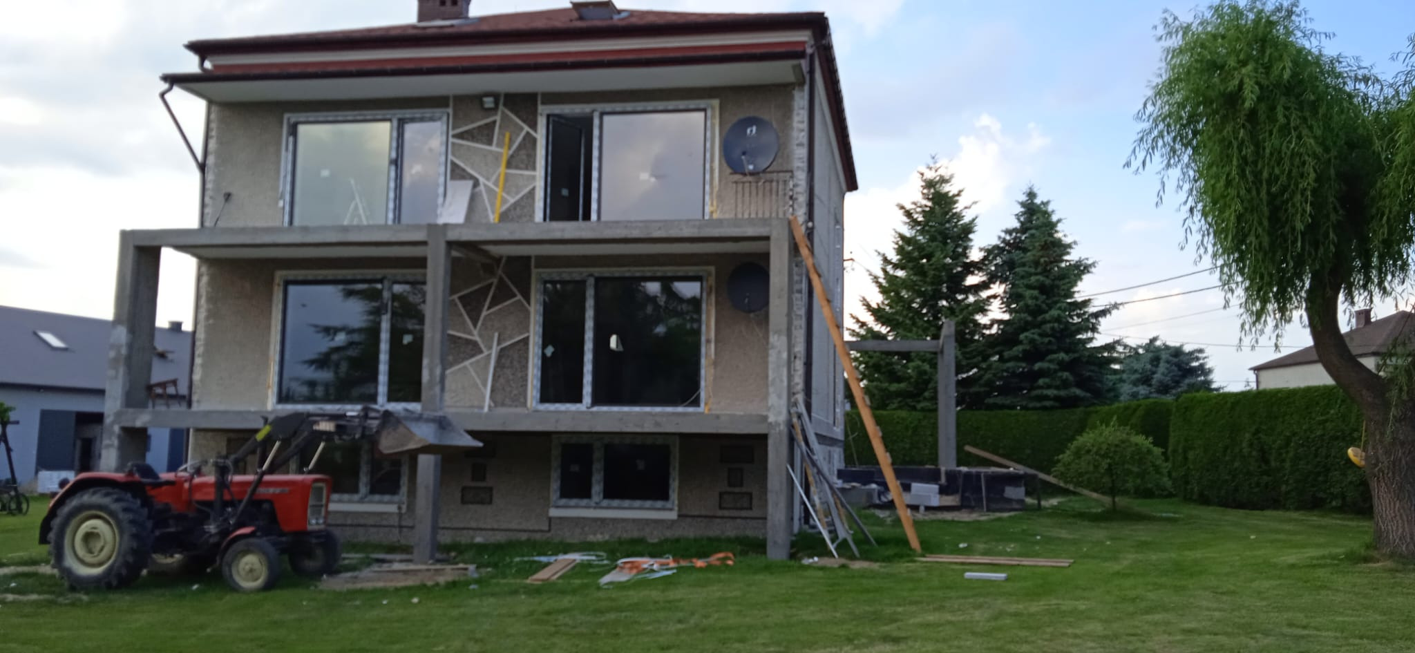 Dom w trakcie budowy z zamontowanymi oknami i traktorem na trawniku, widoczne rusztowania i materiały budowlane.