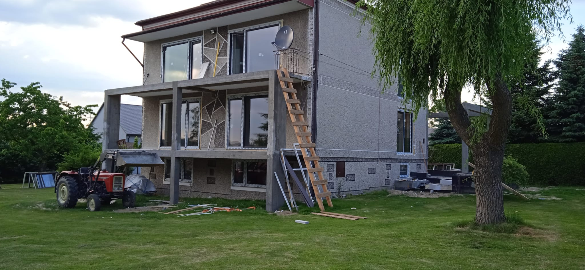 Dwupiętrowy dom w trakcie montażu okien PCV z szarą elewacją, widoczne rusztowanie, drabina, traktor i materiały budowlane.