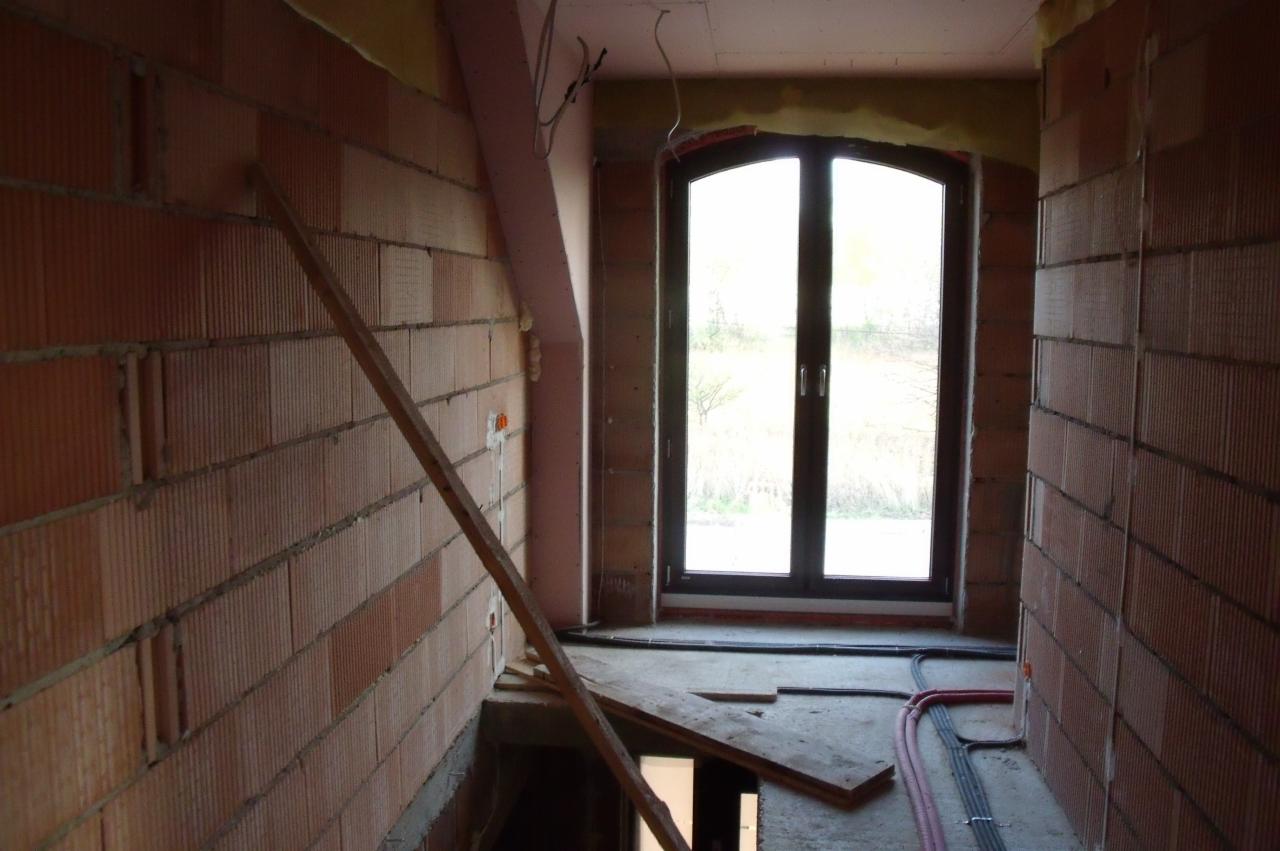 Surowy stan budynku w Kocmyrzowie: widoczne przewody elektryczne w peszlach na podłodze, ściany z cegieł i okno. Etap prac instalacyjnych przed wykończeniem.