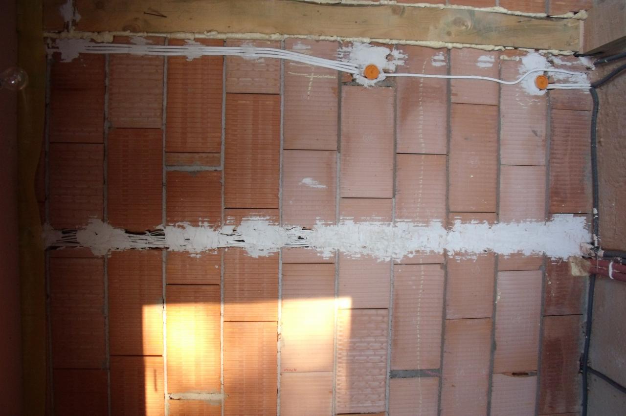 Instalacja elektryczna w trakcie montażu: przewody w białej listwie na ścianie z cegły, widoczne pomarańczowe puszki elektryczne. Etap prac przed tynkowaniem.