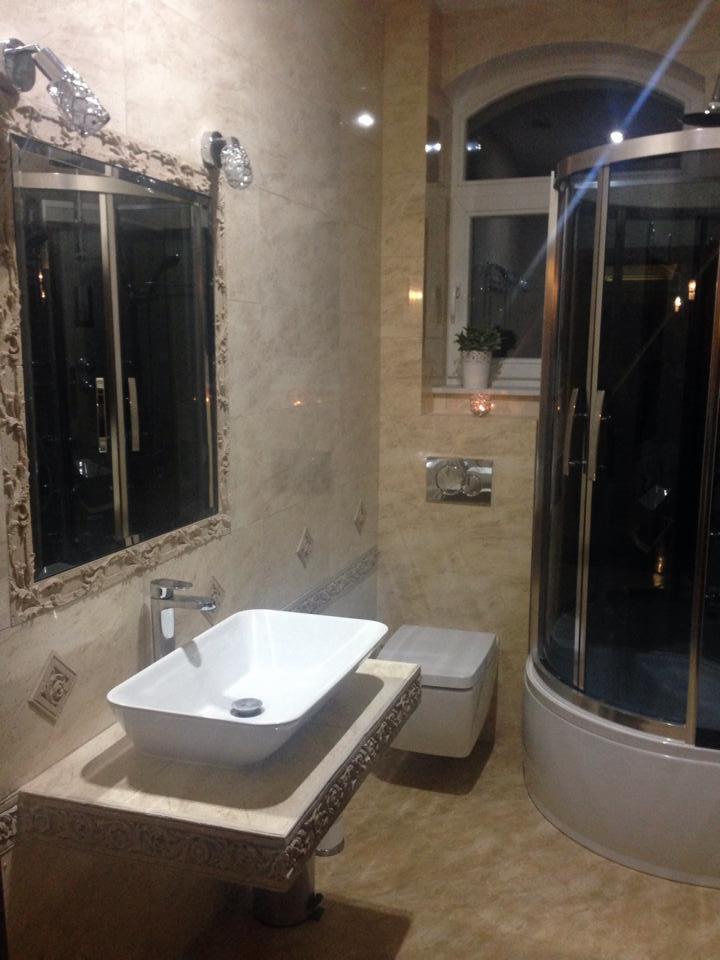 Elegancka łazienka z beżowymi płytkami, kabiną prysznicową z ciemnego szkła, umywalką na zdobionym blacie i lustrem w bogato rzeźbionej ramie.