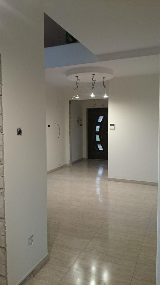 Jasne wnętrze po remoncie: kremowe ściany, błyszcząca podłoga z płytek, nowoczesne oświetlenie sufitowe i ciemne drzwi wejściowe z przeszkleniami.