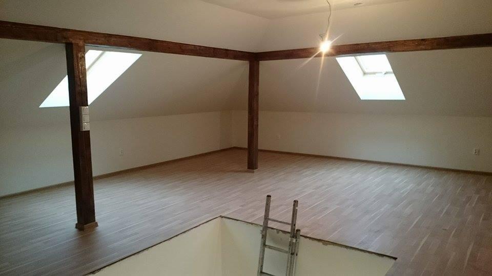 Jasne poddasze po remoncie z drewnianymi belkami, oknami dachowymi i nową podłogą. W tle widoczna drabina prowadząca do otworu w podłodze.