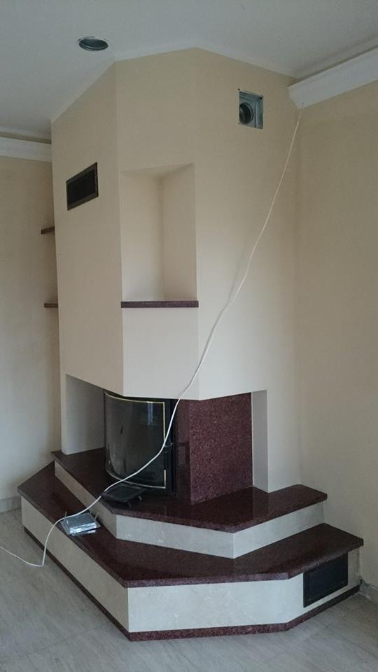 Wykończony kominek z czerwonego granitu i beżowych ścian, z półkami po lewej stronie i kratką wentylacyjną u góry. Widoczny fragment podłogi i kabla.