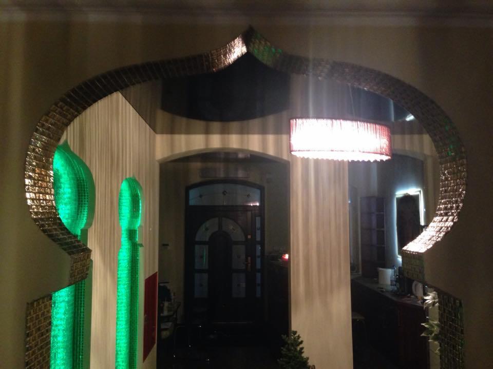 Aranżacja wnętrza z dekoracyjnym łukiem z mozaiki i zielonymi elementami podświetlanymi LED, widok na korytarz z lampą sufitową.