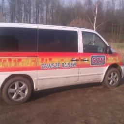 Biały bus z grafiką miejską i napisem 'Zawsze Busem' na boku, zaparkowany na polnej drodze. Widoczne ciemne szyby i numer 525.