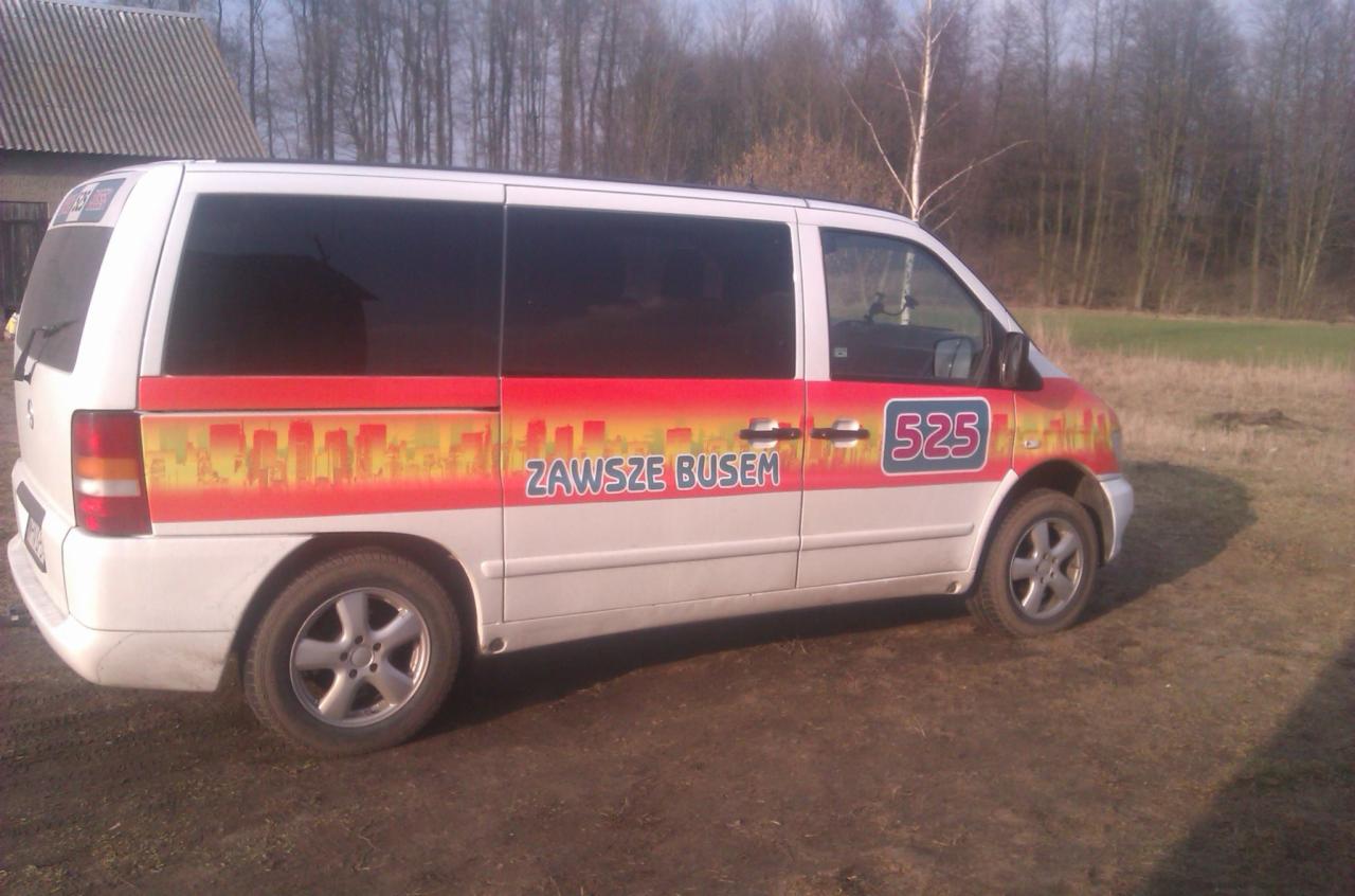 Biały bus z grafiką miejską i napisem 'Zawsze Busem' na boku, zaparkowany na polnej drodze. Widoczne ciemne szyby i numer 525.