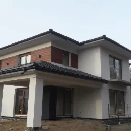 Dwukondygnacyjny dom z białym tynkiem i elementami z drewnianych paneli, widoczny w trakcie wykończenia, z niezabudowaną przestrzenią pod dachem i elementami szalunk&oacute;w w tle.