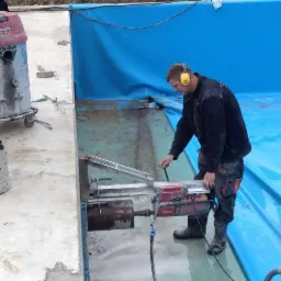 Pracownik w słuchawkach tnie betonową posadzkę wewnątrz niebieskiego basenu, obok odkurzacz przemysłowy i rękawice robocze. Widok z góry, oświetlenie dzienne.