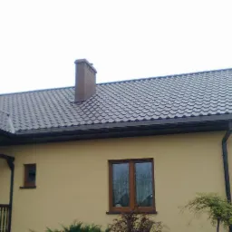 Dom z nową, brązową elewacją i dachem z blachodachówki w odcieniach szarości. Widoczny komin, okna z brązowymi ramami i fragment ganku z balustradą.