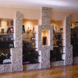 Wnęki ścienne z kamienną okładziną i szklanymi półkami, oświetlone punktowo, wypełnione książkami i dekoracjami. Parkiet na podłodze.