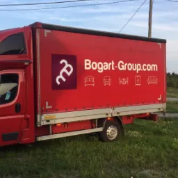 Czerwony samochód dostawczy z logo Bogart-Group.com i ikonami mebli, stojący na trawiastym terenie z słupem elektrycznym w tle.