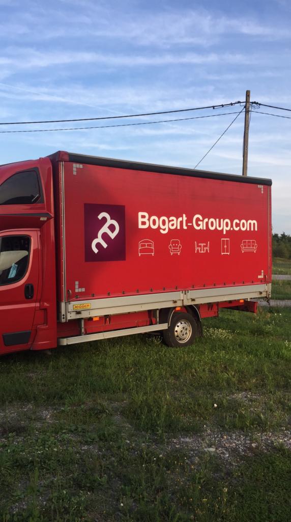 Czerwony samochód dostawczy z logo Bogart-Group.com i ikonami mebli, stojący na trawiastym terenie z słupem elektrycznym w tle.