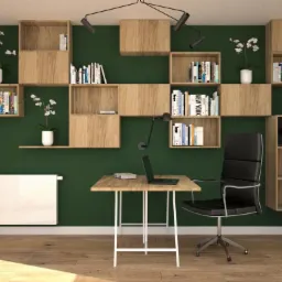 Nowoczesne biuro domowe z zieloną ścianą, regałami z jasnego drewna, biurkiem z laptopem i czarnym krzesłem biurowym na kółkach. Oświetlenie punktowe na suficie i biały grzejnik pod oknem.
