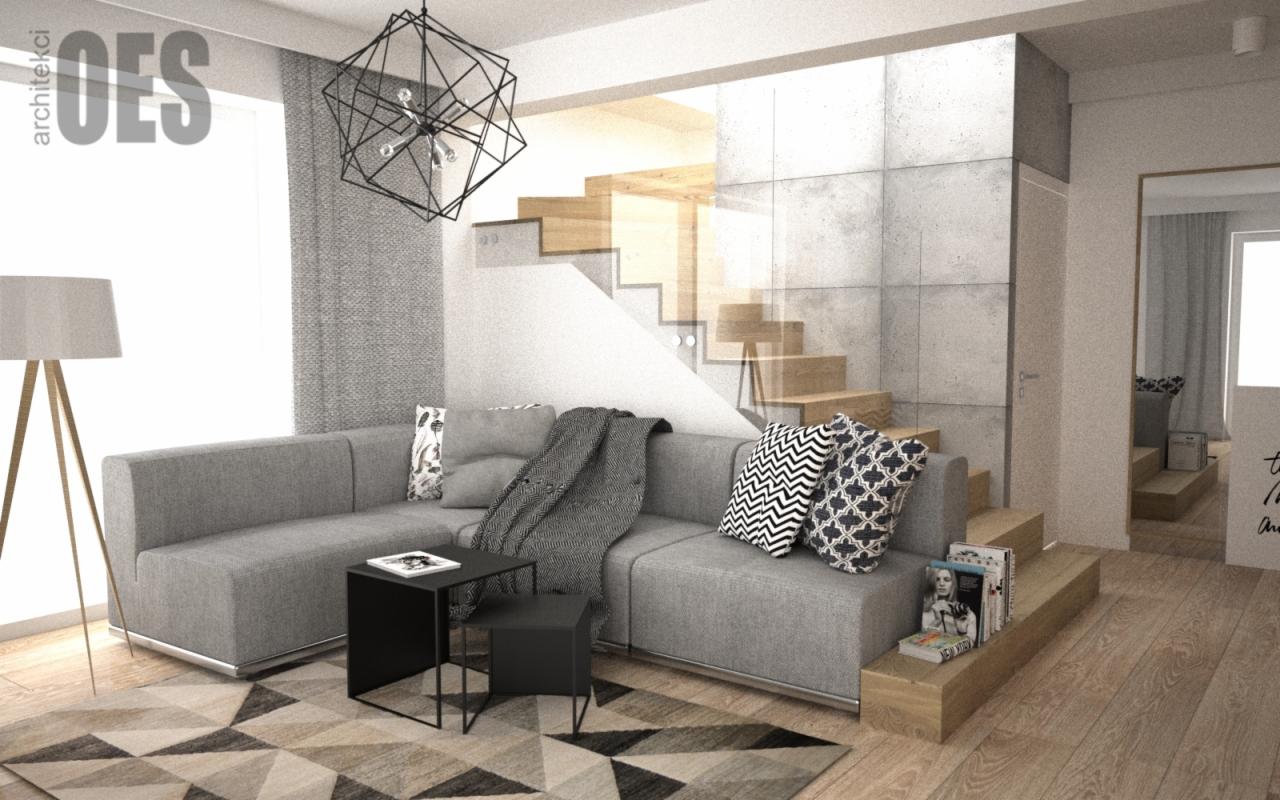 Szary narożnik z geometrycznymi poduszkami i pledem, czarny stolik kawowy, lampa na trójnogu, schody z drewnianymi stopniami i szklaną balustradą, betonowa ściana w salonie z dywanem w geometryczne...