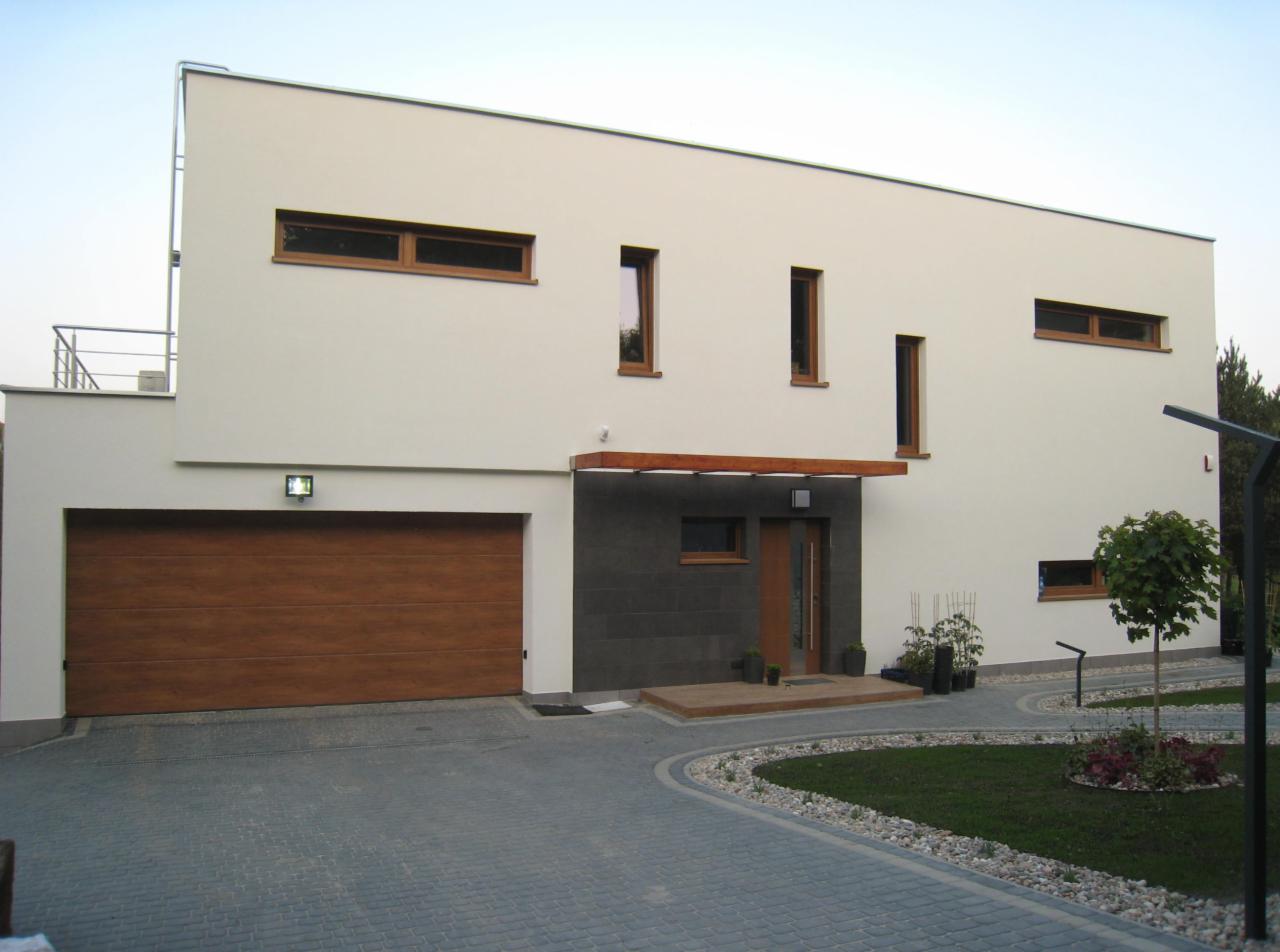 Nowoczesny dom jednorodzinny z płaskim dachem, białymi ścianami, drewnianą bramą garażową i kamienną elewacją przy wejściu, otoczony kostką brukową i trawnikiem z nasadzeniami.