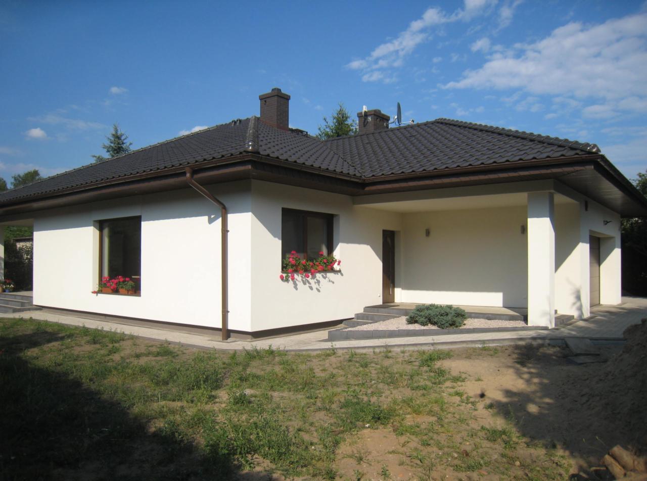 Nowoczesny, parterowy dom jednorodzinny z ciemną dachówką i jasną elewacją, z oknami ozdobionymi czerwonymi pelargoniami w skrzynkach, widok z zewnątrz w słoneczny dzień.