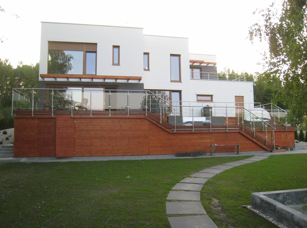 Nowoczesny dom z tarasem na podwyższeniu, drewnianą elewacją i szklanymi balustradami, widok z ogrodu z kamienną ścieżką.
