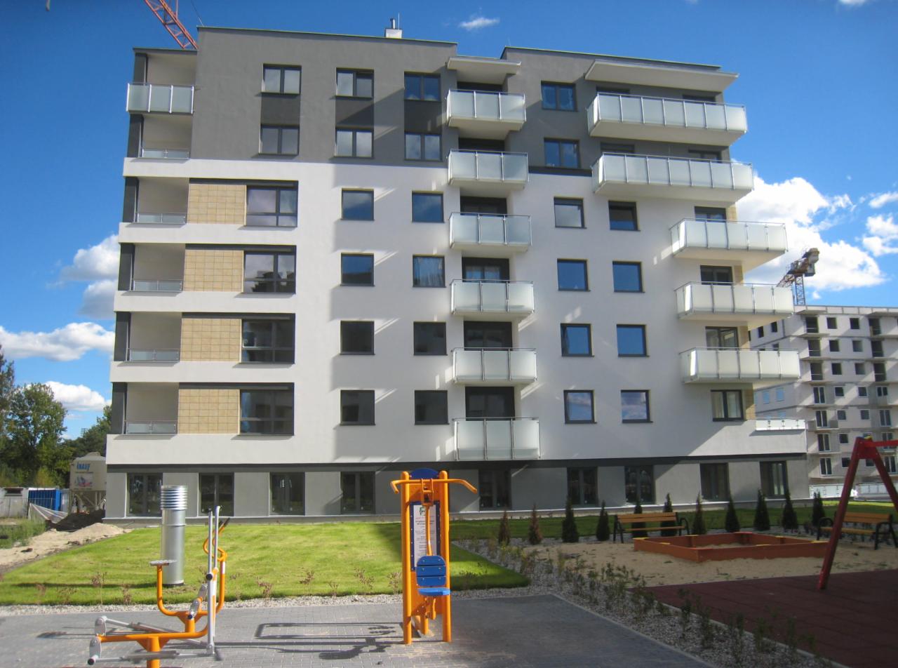 Nowoczesny budynek wielorodzinny z balkonami, plac zabaw z urządzeniami do ćwiczeń na pierwszym planie, oraz żuraw budowlany w tle.