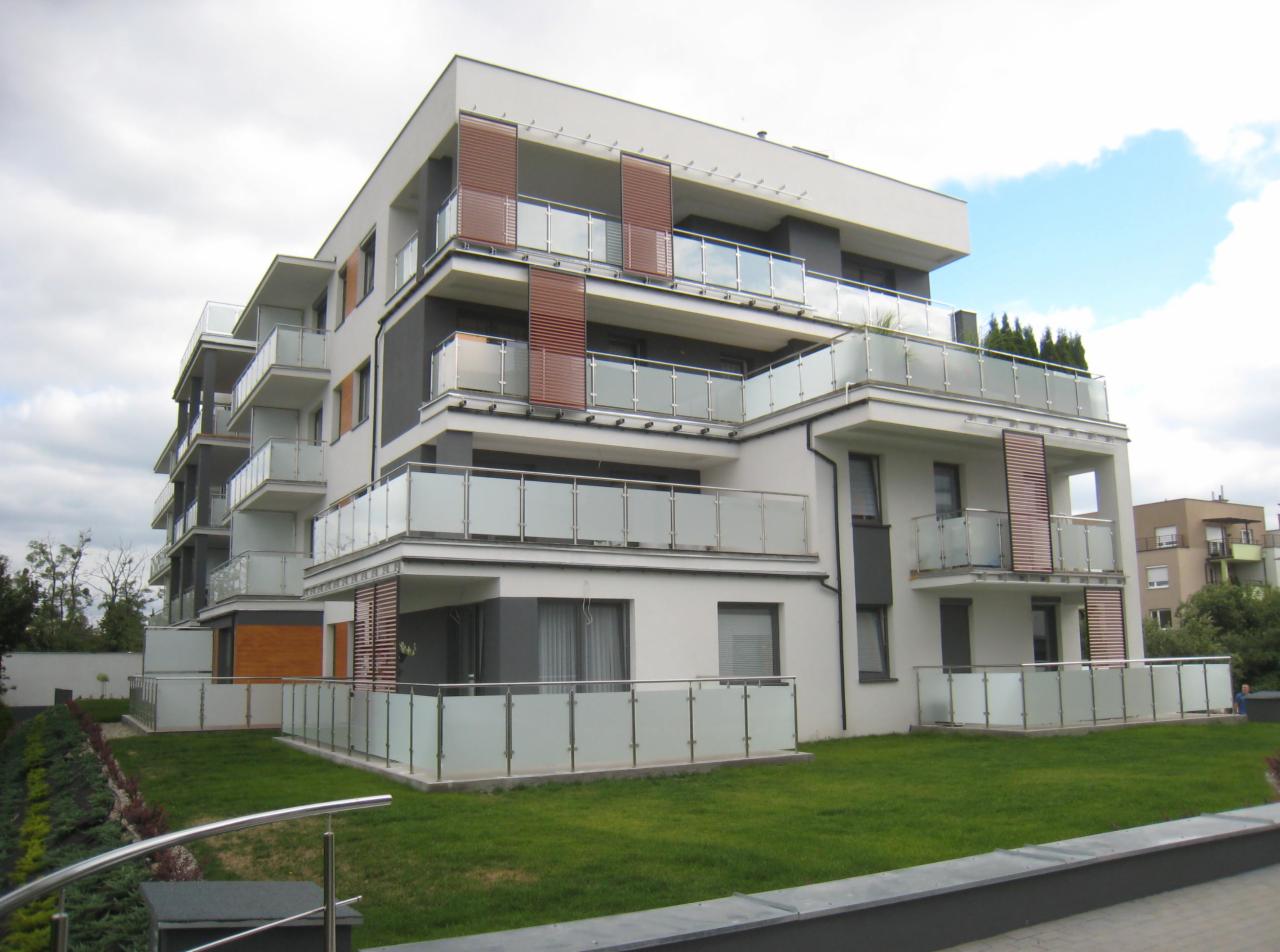 Nowoczesny budynek mieszkalny z balkonami o szklanych balustradach i drewnianymi panelami, widok z poziomu gruntu na tle zachmurzonego nieba.