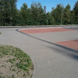 Nowo ułożona, wzorzysta kostka brukowa na parkingu z wyznaczonymi miejscami postojowymi w kolorze czerwonym, obrzeżami z ciemnej kostki i rabatą z młodą zielenią.