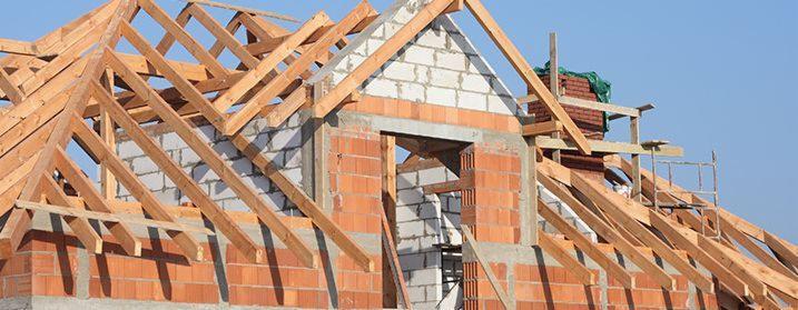 Surowy stan domu w trakcie budowy: czerwona cegła, białe bloczki, drewniana konstrukcja dachu na tle błękitnego nieba. Widoczny komin z zieloną plandeką i rusztowanie.