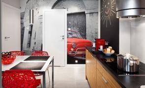 Nowoczesna kuchnia z drewnianymi szafkami, czarnym blatem i płytą indukcyjną. Na ścianie fototapeta z czerwonym Fiatem 500. Czerwone krzesła i owoce na stole dodają koloru.