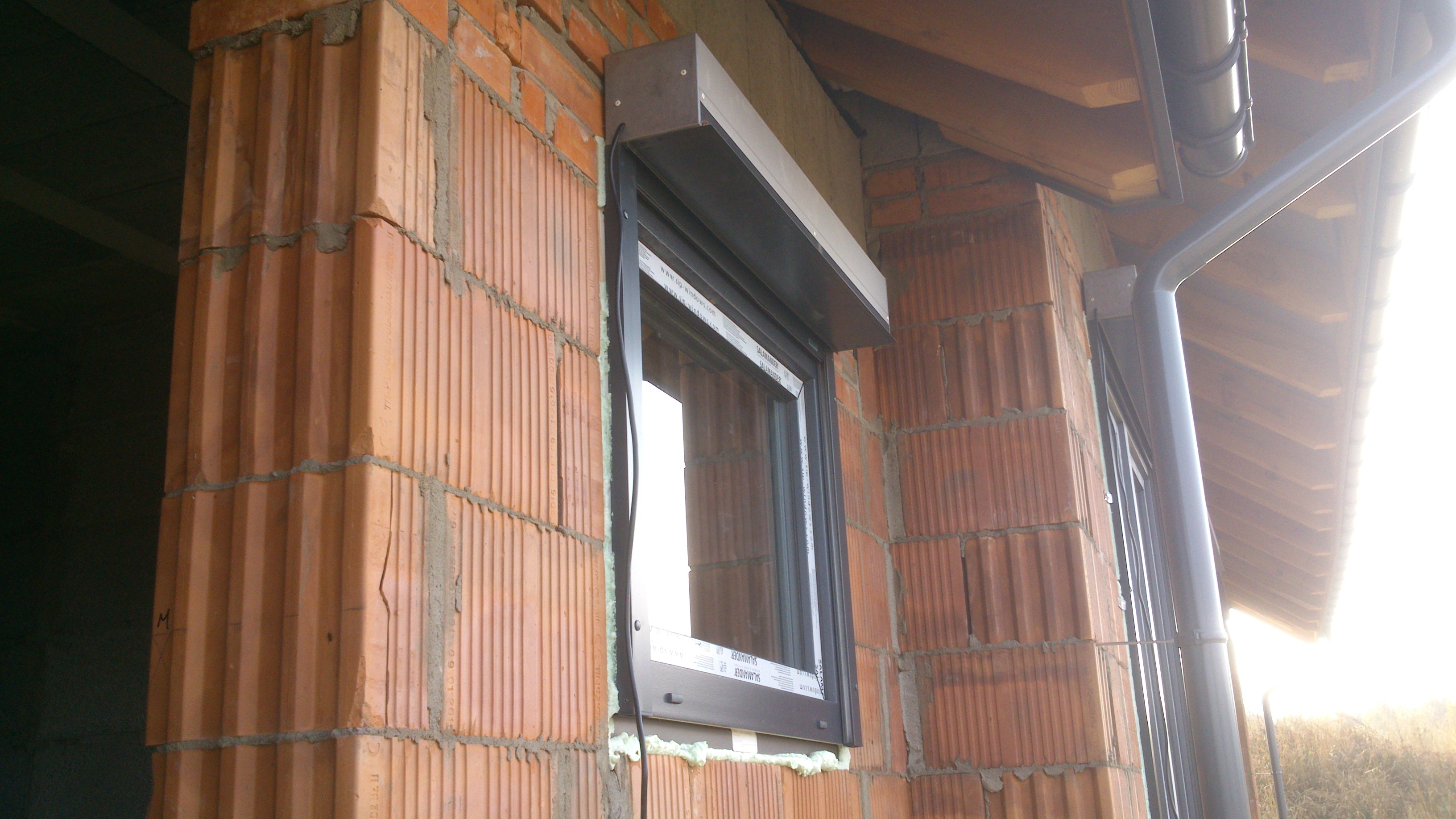 Ceglany budynek z oknem PCV i roletą zewnętrzną w trakcie montażu. Widoczna piana montażowa i surowe wykończenie muru.